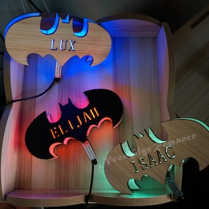 May include: Three wooden Batman-shaped signs with LED lights. The signs are illuminated in blue, red, and green. The names "LUX", "ELIJAH", and "ISAAC" are engraved on the signs.