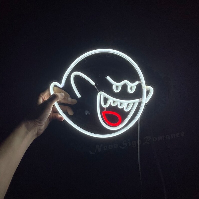 May include: White neon sign shaped like a ghost with a mischievous grin and a red mouth. The sign is lit up and is being held by a hand.