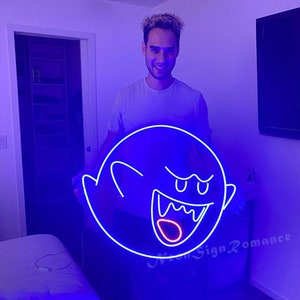 May include: A person holds a blue neon sign shaped like a ghost with a red mouth and white eyes. The sign is lit up and glowing.