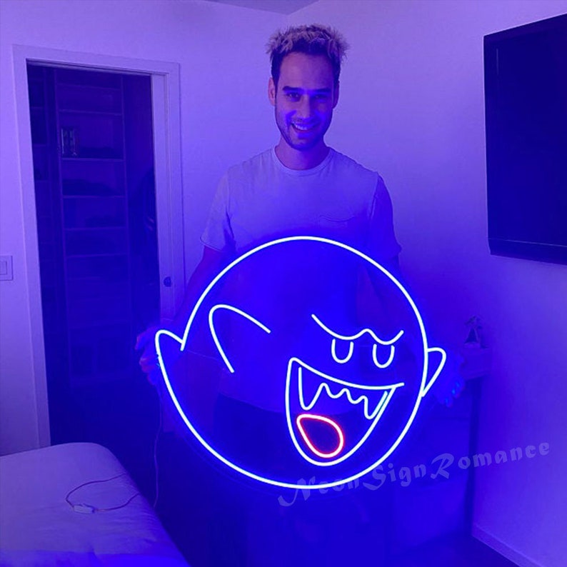 May include: A person holds a blue neon sign shaped like a ghost with a red mouth and white eyes. The sign is lit up and glowing.