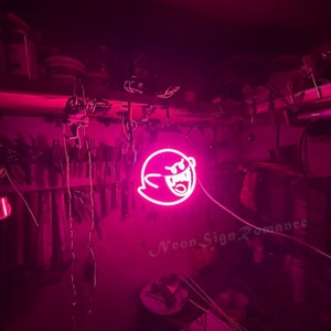 May include: Pink neon sign of a ghost with a toothy grin. The sign is hanging from the ceiling in a dark room. The text "Neon Sign Romance" is visible in the bottom right corner of the image.