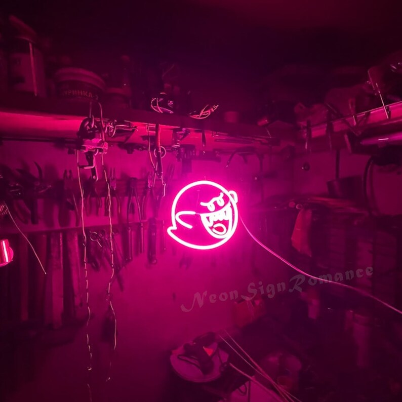 May include: Pink neon sign of a ghost with a toothy grin. The sign is hanging from the ceiling in a dark room. The text "Neon Sign Romance" is visible in the bottom right corner of the image.