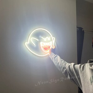 May include: White neon sign of a ghost with a wide open mouth and teeth. The ghost is lit up with a bright white light. The sign is mounted on a white wall.