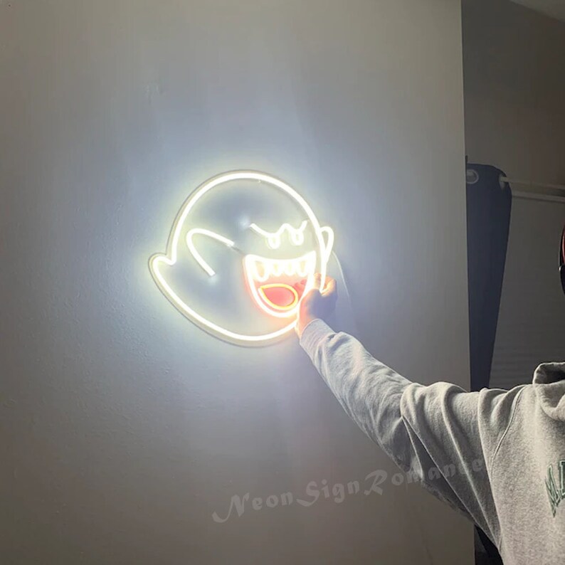 May include: White neon sign of a ghost with a wide open mouth and teeth. The ghost is lit up with a bright white light. The sign is mounted on a white wall.