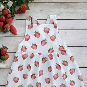 May include: A white sleeveless dress with a strawberry print. The dress has a gathered skirt and adjustable straps. The dress has a small label that says "Teorya".