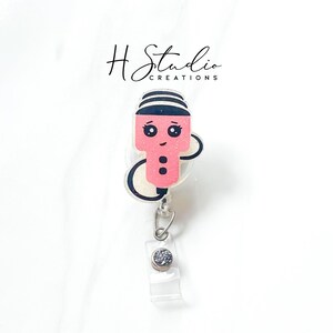 May include: A pink and black snowman-themed badge reel with a clear plastic strap and a silver-coloured clip. The snowman has a striped hat, black eyes, and a smiling face. The badge reel is attached to a clear plastic retractable cord.