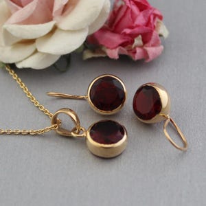 May include: A gold jewellery set consisting of a pendant on a delicate chain and two matching earrings. Each piece features a round, deep red garnet gemstone set in a smooth gold bezel. The earrings have curved wire hooks for secure attachment.