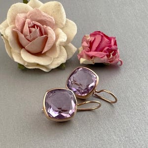 Può includere: Un paio di orecchini color oro con pietre quadrate viola chiaro. Gli orecchini sono su uno sfondo grigio, con una grande rosa rosa chiaro e crema e una rosa più piccola rosa scuro sullo sfondo.