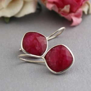 Könnte beinhalten: Ein Paar silberne Ohrhänger mit facettierten, quadratischen, tiefroten Edelsteinen. Die Ohrringe haben ein schlichtes Design, wobei die Edelsteine in einer silbernen Lünette gefasst sind.