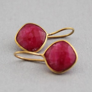 May include: A pair of gold-toned drop earrings featuring square, faceted, ruby-red gemstones. The earrings have a simple, elegant design with a gold frame around each stone and a curved hook for pierced ears. The earrings are displayed against a neutral background.