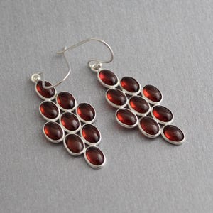 May include: A pair of silver earrings with a cluster of oval, dark red gemstones. The gemstones are set in a silver frame and arranged in a cascading pattern. The earrings have a hook closure.