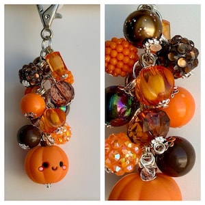 May include: A keychain featuring a cluster of assorted beads in shades of orange, brown, and iridescent tones, attached to a silver-toned metal lobster clasp. The arrangement includes smooth, faceted, and textured beads, finished with a pumpkin charm.