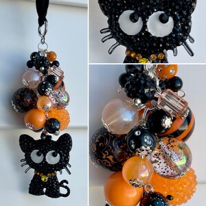 May include: A black cat charm with rhinestone accents, paired with a cluster of beads in black, orange, and iridescent colors. The charm has white eyes and a yellow collar. The beads include various sizes and textures, creating a unique accessory.