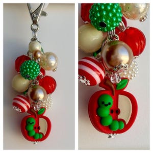 May include: A keychain featuring a silver-toned metal lobster clasp and a cluster of assorted beads in red, green, cream, and metallic gold. The beads include smooth, textured, and striped spheres, finished with a red apple charm with a green caterpillar.