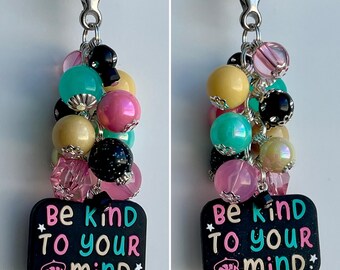 Könnte beinhalten: Ein schwarzer Anhänger mit dem Text "Be kind to your mind" in rosa und türkisfarbener Schrift, mit einem silbernen Schlüsselanhänger und einem Bündel bunter Perlen, darunter rosa, türkis, gelb, schwarz und schillernd.