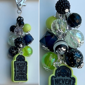 May include: A Halloween-themed charm featuring a tombstone pendant with the text "NEVER TRUST THE LIVING." The charm includes a mix of black, green, and iridescent beads, along with a silver-tone clasp. The pendant is outlined in neon green.