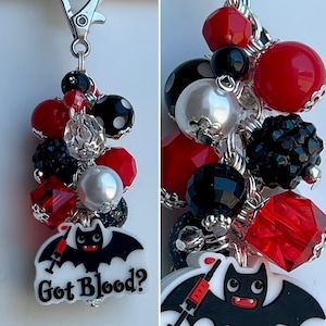 May include: A Halloween-themed charm with a silver clasp. It features a cluster of red, black, and silver beads, including faceted and round shapes. A white charm with a black bat and the text "Got Blood?" is attached.