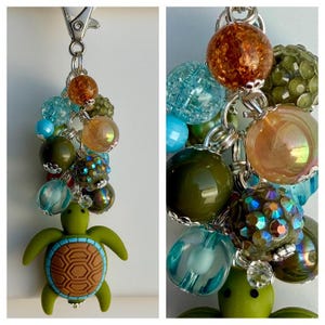 May include: A keychain featuring a green turtle charm attached to a cluster of assorted beads in shades of amber, olive green, and turquoise. The beads include crackled, iridescent, and faceted textures, all connected by silver-toned metal rings.