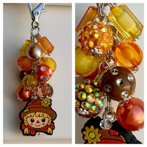 May include: A keychain featuring a collection of orange, yellow, and brown beads in various shapes and textures, including faceted, pearlized, and polka-dot finishes. A flat, cartoon-style scarecrow charm with a yellow sunflower hangs at the bottom.