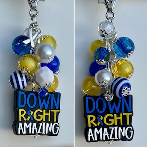 May include: A blue and yellow beaded keychain with a charm that says "Down Right Amazing". The charm is attached to a silver ring with a blue ribbon.