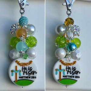 May include: White charm with a green, blue, and brown beaded tassel. The charm has a white background with a brown cross, a blue banner, and the text "He is risen Matthew 28:6".