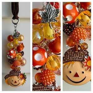 May include: A decorative charm featuring a brown fabric strap, a silver-toned maple leaf charm, and a collection of orange, yellow, and brown beads in various textures. At the base hangs a smiling scarecrow face charm with a brown hat and flower.
