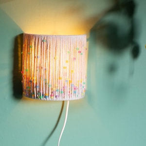 May include: A cylindrical wall lamp with a white shade adorned with vertical strands and colorful beads. The lamp is lit, casting a warm glow against a teal wall. A white cord extends from the bottom.