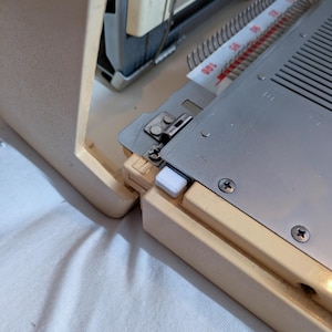 May include: Close-up of a vintage knitting machine. The machine has a cream-colored frame and a silver metal bed with a numbered scale. A white plastic piece is visible on the side. The machine is on a white surface.
