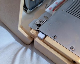 May include: Close-up of a vintage knitting machine. The machine has a cream-coloured frame and a silver metal bed with a numbered scale. A white plastic piece is visible on the side. The machine is on a white surface.