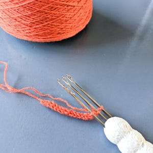 May include: A ball of orange yarn and a white-handled knitting tool with three needles are visible. An orange knitted chain is attached to the needles. The background is a solid blue surface. Knitting and craft supplies.