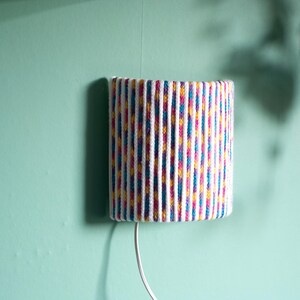 May include: A cylindrical wall-mounted lamp with a colorful striped pattern in white, pink, yellow, and blue. The lamp is attached to a mint green wall with a white cord. The lamp is a decorative lighting fixture.