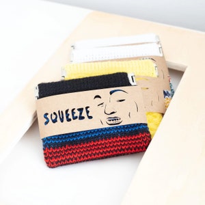 Könnte beinhalten: Ein Stapel bunter Strick-Covers, jedes mit einem einzigartigen Design. Das vordere Cover ist schwarz mit roten und blauen Streifen, und das Etikett trägt die Aufschrift "SQUEEZE" mit einem lächelnden Gesicht. Weitere Covers sind gelb, weiß und gemustert.