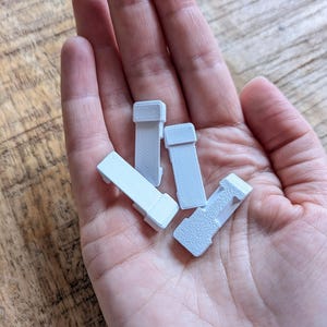 May include: Four small, rectangular, light grey plastic objects with a textured surface are held in a hand. The objects have a rectangular shape with a raised section on one end. The objects are likely small parts or components.