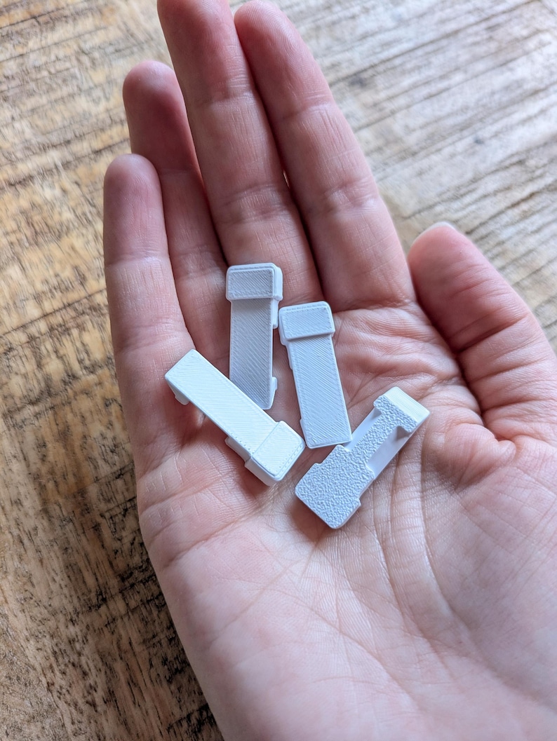 May include: Four small, rectangular, light grey plastic objects with a textured surface are held in a hand. The objects have a rectangular shape with a raised section on one end. The objects are likely small parts or components.