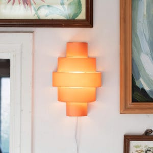 Op de afbeelding: Een oranje wandlamp met een gelaagd, geometrisch ontwerp. De lamp is gemonteerd op een witte muur, met een wit snoer. Kunstwerken in houten frames omringen de lamp.