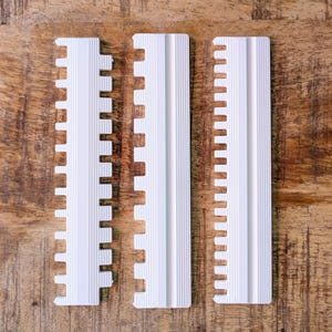 Puede incluir: Tres peines de plástico blanco con un diseño acanalado, dispuestos sobre una superficie de madera. Los peines tienen una serie de dientes a lo largo de un borde y una superficie ranurada en el otro. Los objetos están espaciados y parecen nuevos.