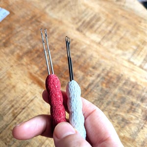 May include: Two crochet hooks with textured handles. One hook has a red handle, the other has a white handle. The metal hook portion is silver and black. The hooks are designed for yarn crafts and fiber art projects.