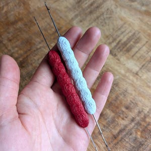 May include: Two crochet hooks with textured handles. One hook has a red handle, and the other has a light blue handle. The hooks are held in a hand, with the wooden surface visible in the background. The hooks are used for crochet and other crafts.