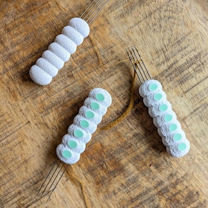 May include: Three white yarn tools with metal tines and textured handles. One tool has a solid white handle, while the other two have handles with light green accents. The tools are arranged on a wooden surface.