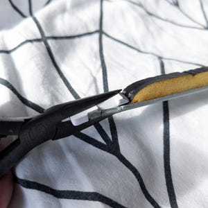 May include: Close-up of black pliers cutting a section of a black and gold object. The object has a metallic frame and a padded, textured surface. The background is a white fabric with a black tree branch pattern.