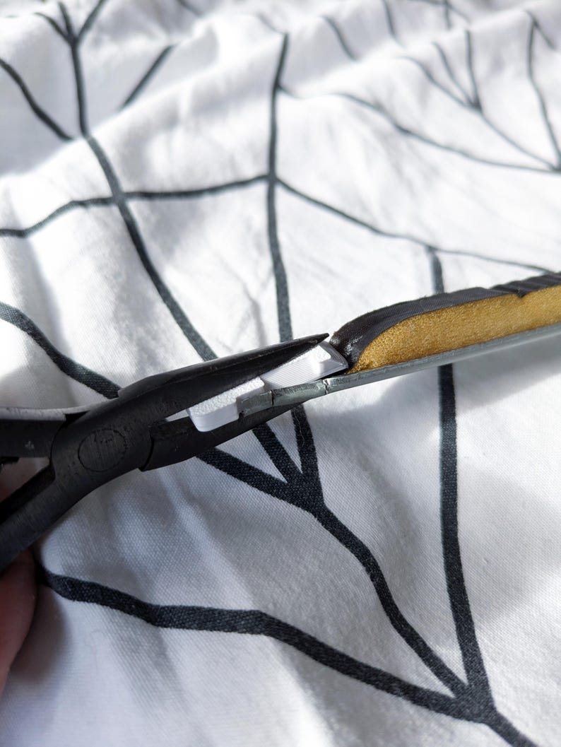 May include: Close-up of black pliers cutting a section of a black and gold object. The object has a metallic frame and a padded, textured surface. The background is a white fabric with a black tree branch pattern.