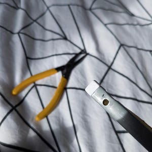 May include: A pair of yellow-handled pliers and a silver and black tool with a white tip are shown. The tools are on a white patterned surface.