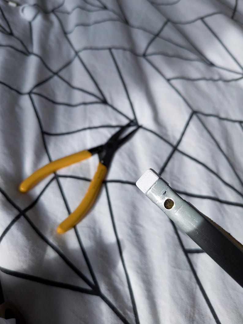 May include: A pair of yellow-handled pliers and a silver and black tool with a white tip are shown. The tools are on a white patterned surface.
