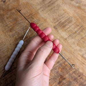 May include: A red needle felting tool with a textured handle is held in a hand. A second, similar tool with a gray handle is in the background. The tools are designed for fiber art and crafting.