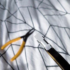 May include: A black and gold belt with a white metal buckle. Yellow and black pliers are in the background. The items are on a white patterned surface.