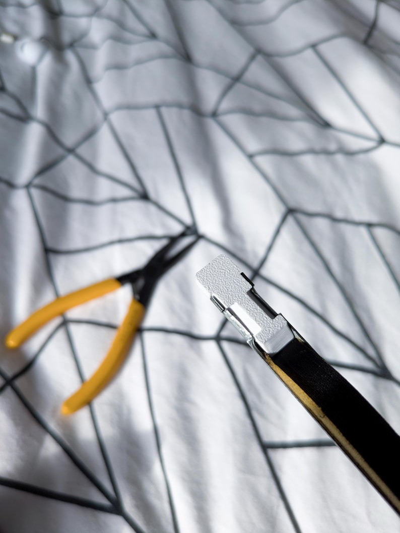 May include: A black and gold belt with a white metal buckle. Yellow and black pliers are in the background. The items are on a white patterned surface.