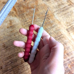 Puede incluir: Dos ganchillos con varillas metálicas y mangos texturizados. Un mango es rojo, el otro azul claro. Los ganchillos se sostienen en una mano sobre un fondo de madera. Una regla es visible en el fondo.