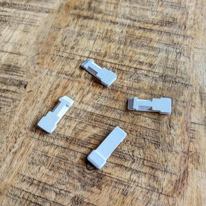 May include: Four small, light grey plastic pieces with a rectangular shape and a T-shaped end are scattered on a wooden surface. The pieces appear to be components of a larger item, possibly a furniture part or a small appliance.