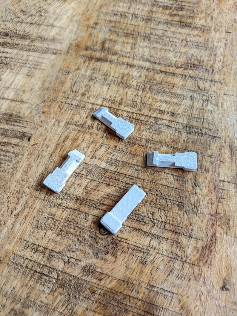 May include: Four small, light grey plastic pieces with a rectangular shape and a T-shaped end are scattered on a wooden surface. The pieces appear to be components of a larger item, possibly a furniture part or a small appliance.