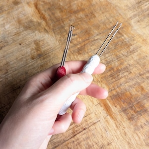 May include: Two handmade hair tools are held against a wooden background. One tool has a red, round handle, and the other has a white, textured handle. Both tools have thin, silver metal prongs.